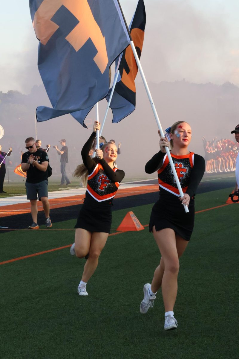 Running with flags, senior Audrina Becker and Evie Metcalfe perform at the football game. On Sept. 12, the cheer team and student section dressed in red, white and blue to honor heroes night. 